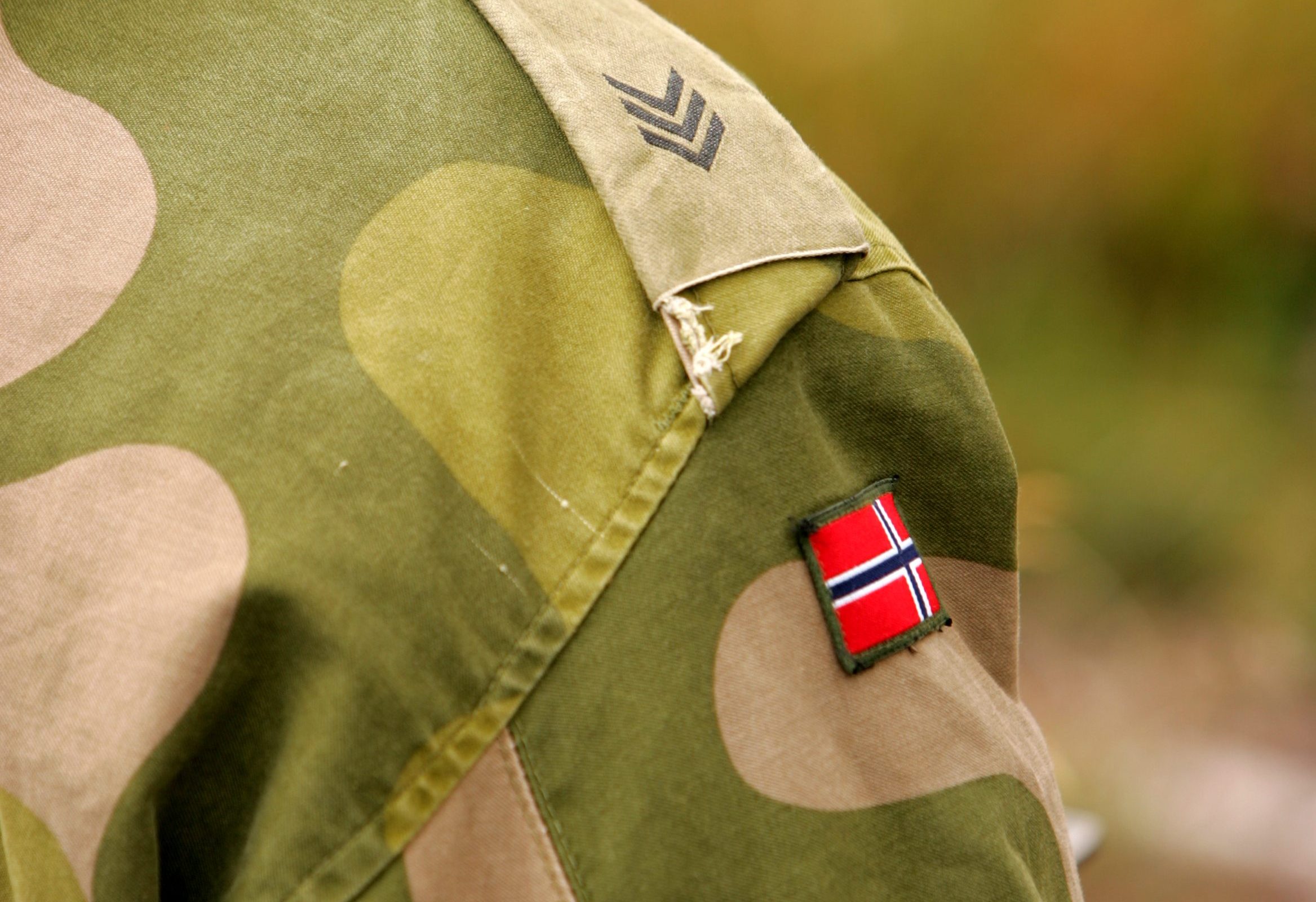 Nærbilde av en norsk sersjants venstre skulder i Forsvaret: olivengrønt uniformstøy med tre nedovervendte chevroner som angir sersjantgrad, og et lite rektangulært norsk flagg sydd under rangstripene.