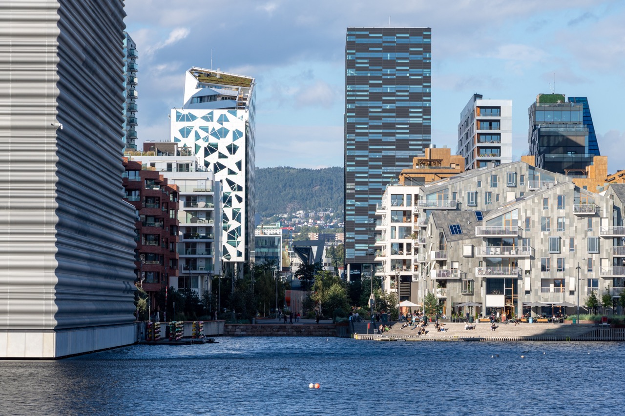 Foto fra 2024 av Bjørvika i Oslo viser et moderne bylandskap med innovative bygninger i bølgende og geometriske former. Foran i bildet skimtes rolig vann og en liten strand der folk nyter området. I bakgrunnen reiser grønne åser seg, og skaper en kontrast til den moderne arkitekturen – et bilde på Oslos satsing på samspill mellom natur og byutvikling.