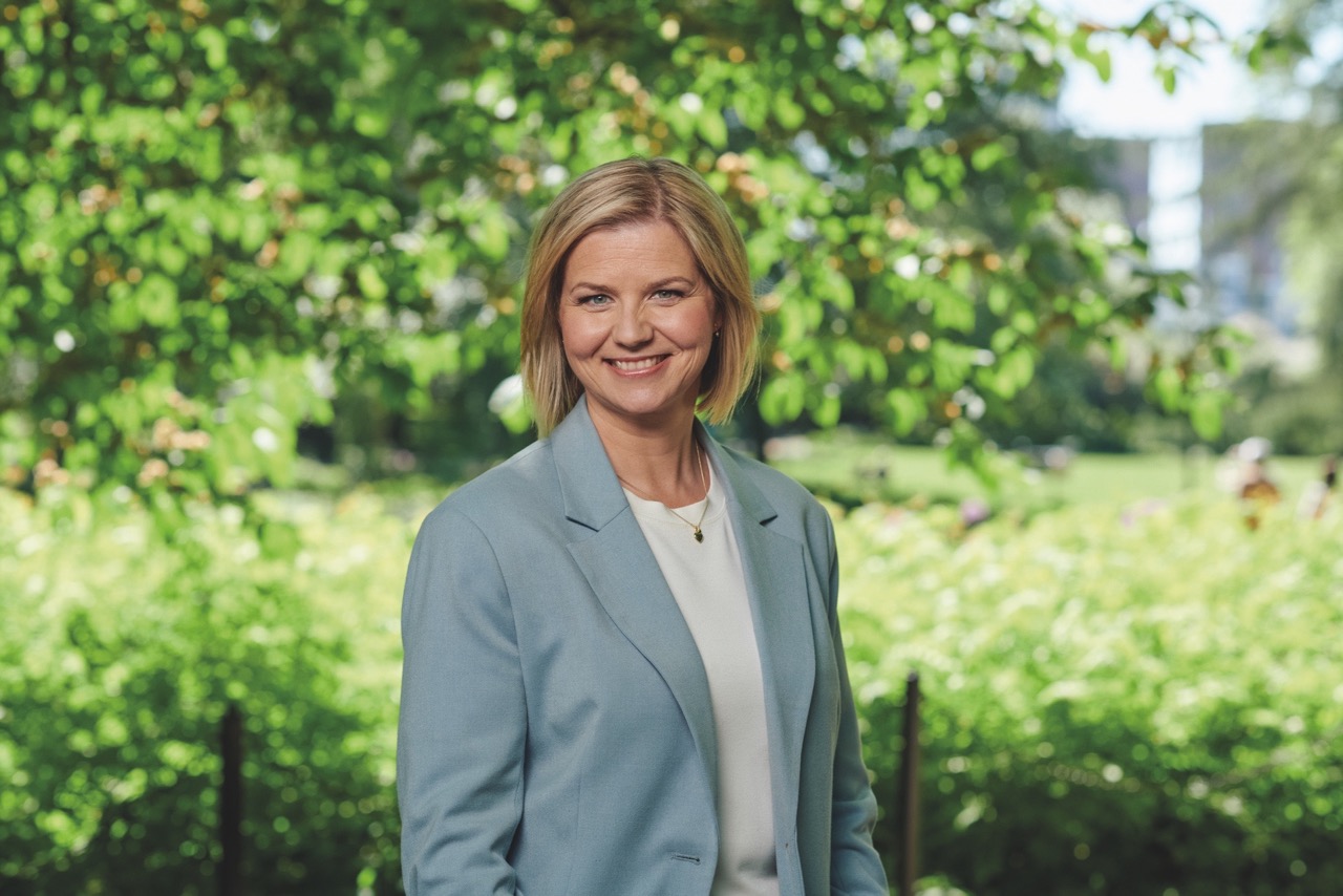 Portrett av en smilende, Guri Melby iført en lys blå blazer og hvit bluse, stående foran frodig, grønn løvverk i en solfylt park.