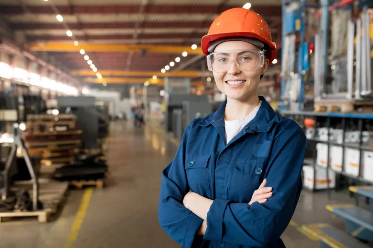 Ung kvinnelig tekniker i blå arbeidsuniform, vernebriller og gul sikkerhetshjelm, smilende.