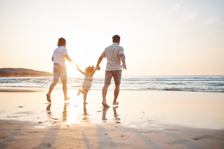 To menn som holder hender med sin lille datter mens de går langs en solfylt strand. Det varme sollyset og det klare havet gir en følelse av trygghet, kjærlighet og likestilling.