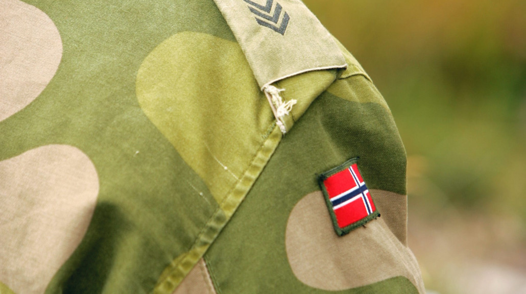 Nærbilde av en norsk sersjants venstre skulder i Forsvaret: olivengrønt uniformstøy med tre nedovervendte chevroner som angir sersjantgrad, og et lite rektangulært norsk flagg sydd under rangstripene.