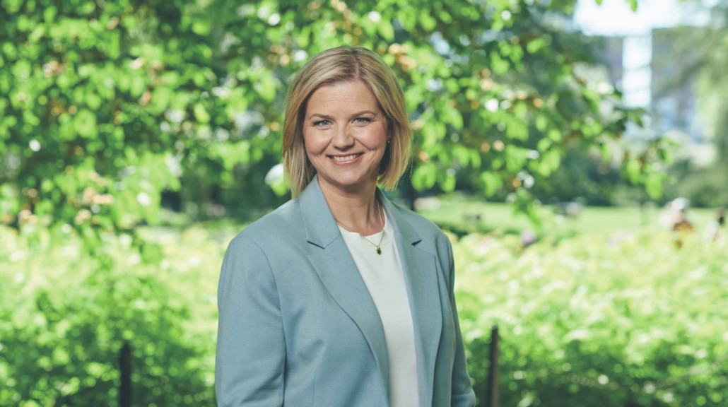 Portrett av en smilende, Guri Melby iført en lys blå blazer og hvit bluse, stående foran frodig, grønn løvverk i en solfylt park.