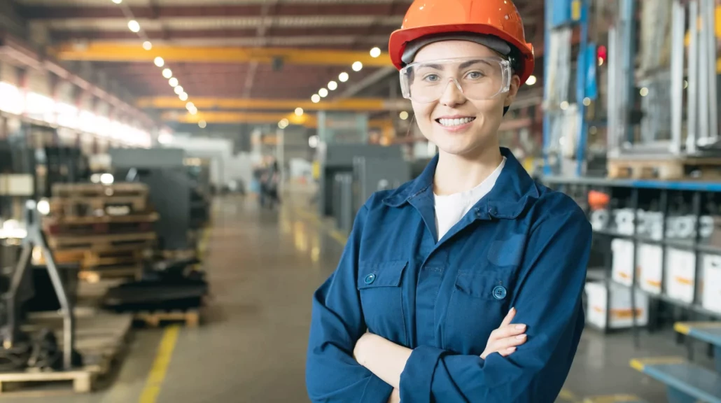 Ung kvinnelig tekniker i blå arbeidsuniform, vernebriller og gul sikkerhetshjelm, smilende.