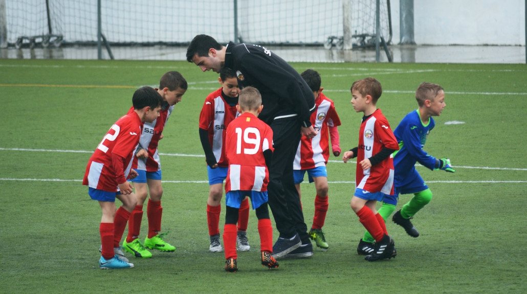 Barn i roede og blaa fotballdrakter staar samlet paa kunstgress i et hall med treneren i midten.