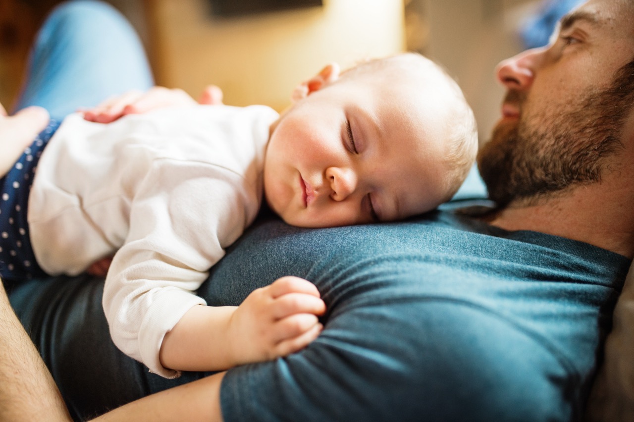 Far og babyjente sover sammen på sofaen – et nært øyeblikk som viser verdien av pappaperm og tid med familien.