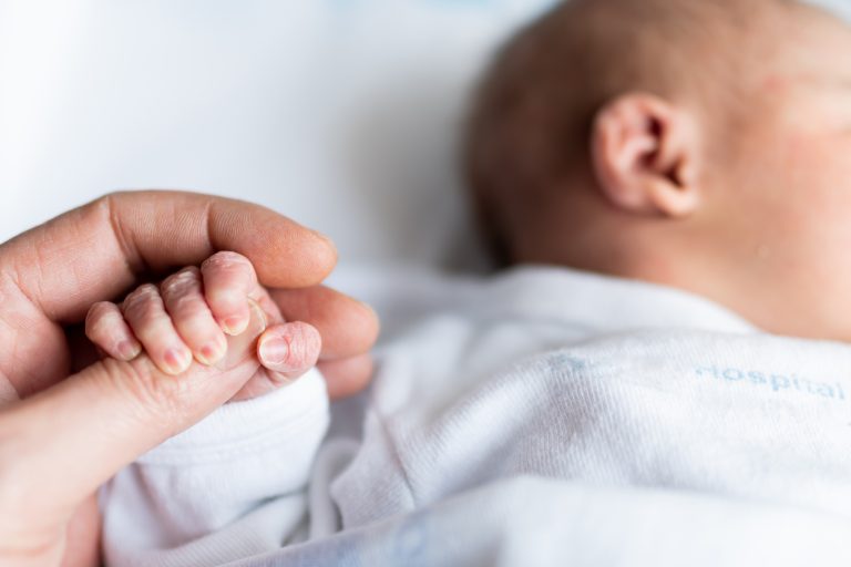En nyfødt baby holder rundt fingeren til en voksen hånd. Bildet symboliserer trygghet, omsorg og behovet for en styrket fødselsomsorg og bedre støtte til foreldre og barn i helsetjenesten.