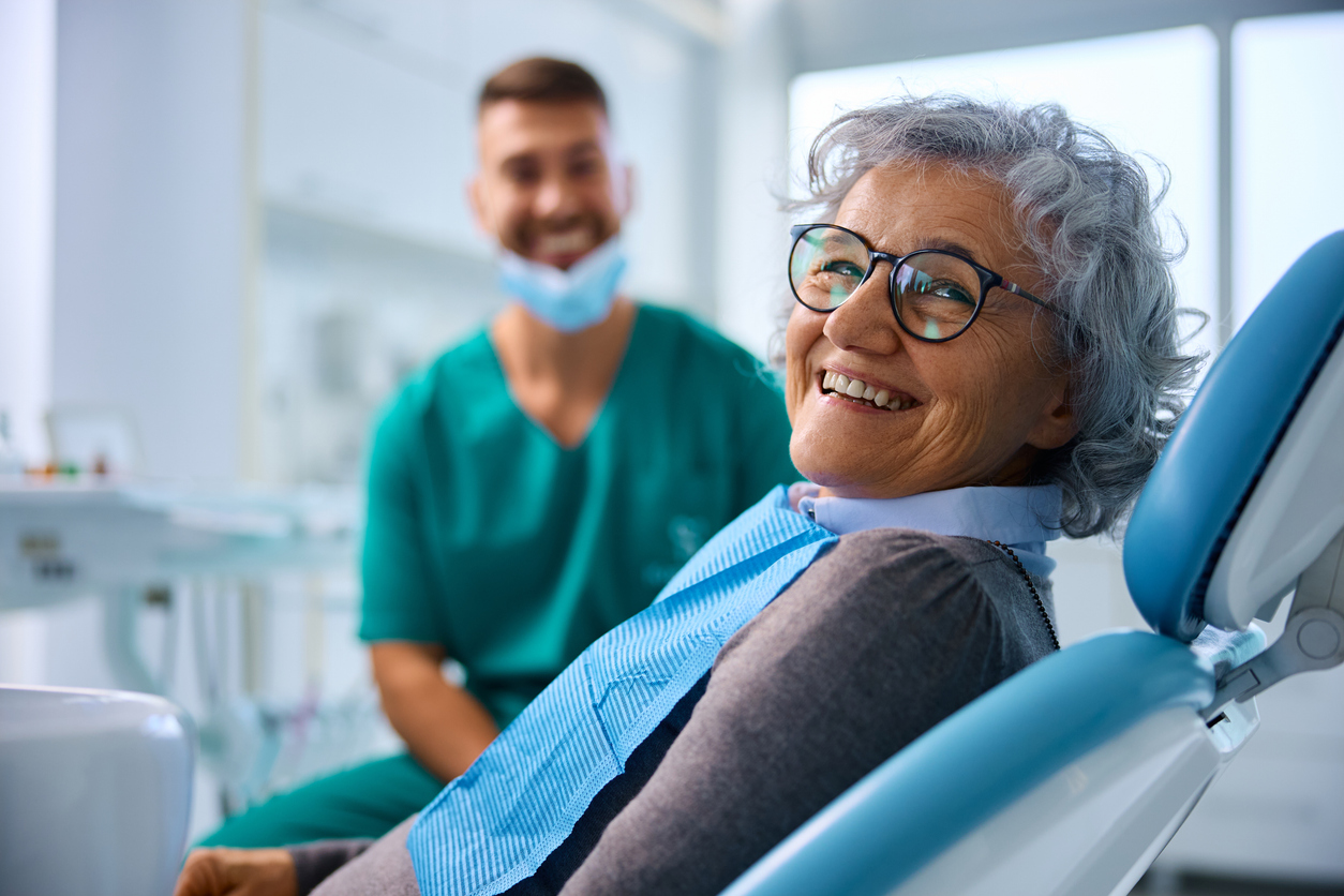 Glad eldre kvinne på tannklinikk ser i kameraet, med tannlegen i bakgrunnen.