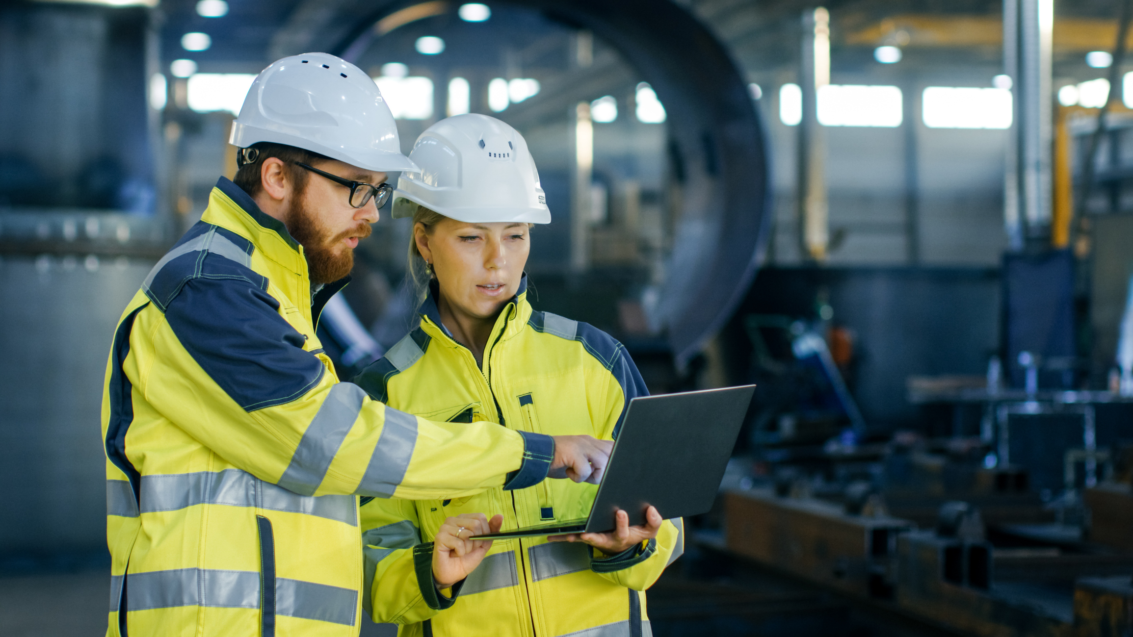 En kvinnelig og en mannlig industriingeniør med vernehjelm diskuterer et nytt prosjekt mens de ser på en bærbar PC i en produksjonshall