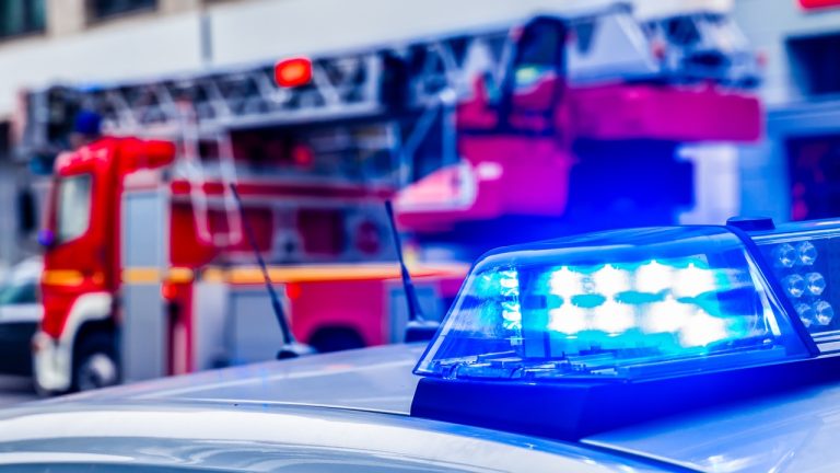 Nærbilde av et blått, blinkende taklys på en politibil i forgrunnen, med en rød brannbil synlig i bakgrunnen – en visuell framstilling av politi- og brannvesenets samarbeid innen rettssikkerhet og beredskap