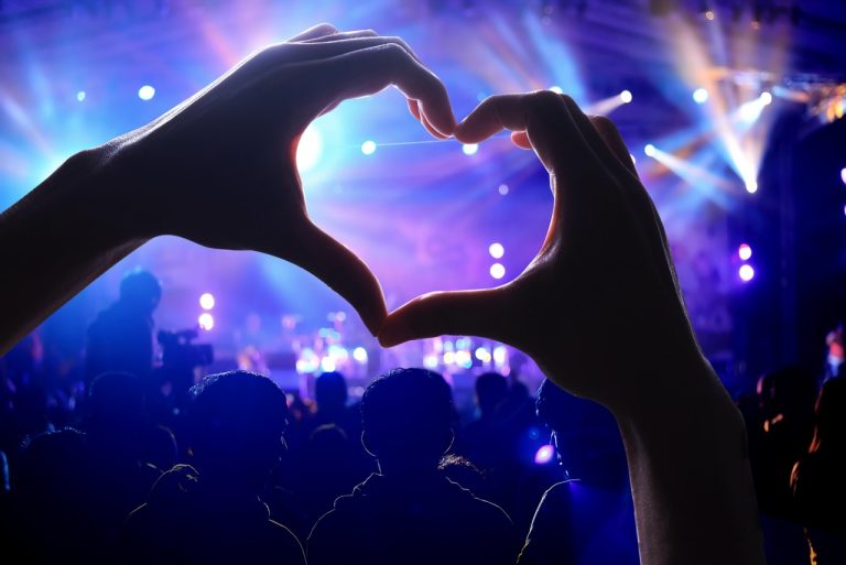 Silhuetter av et konsertpublikum med opphøyde armer mot en fargerik scene, hvor to hender i forgrunnen danner et hjerte; symboliserer fellesskap, kulturglede og frivillig engasjement
