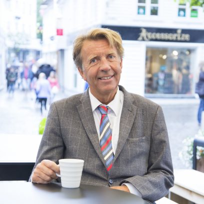 Portrettbilde av Per A. Thorbjørnsen,, sittende ved et kafébord og holder en kaffekopp  Han ser framover med et lyttende uttrykk, omgitt av et dempet urbant kaféinteriør i bakgrunnen.