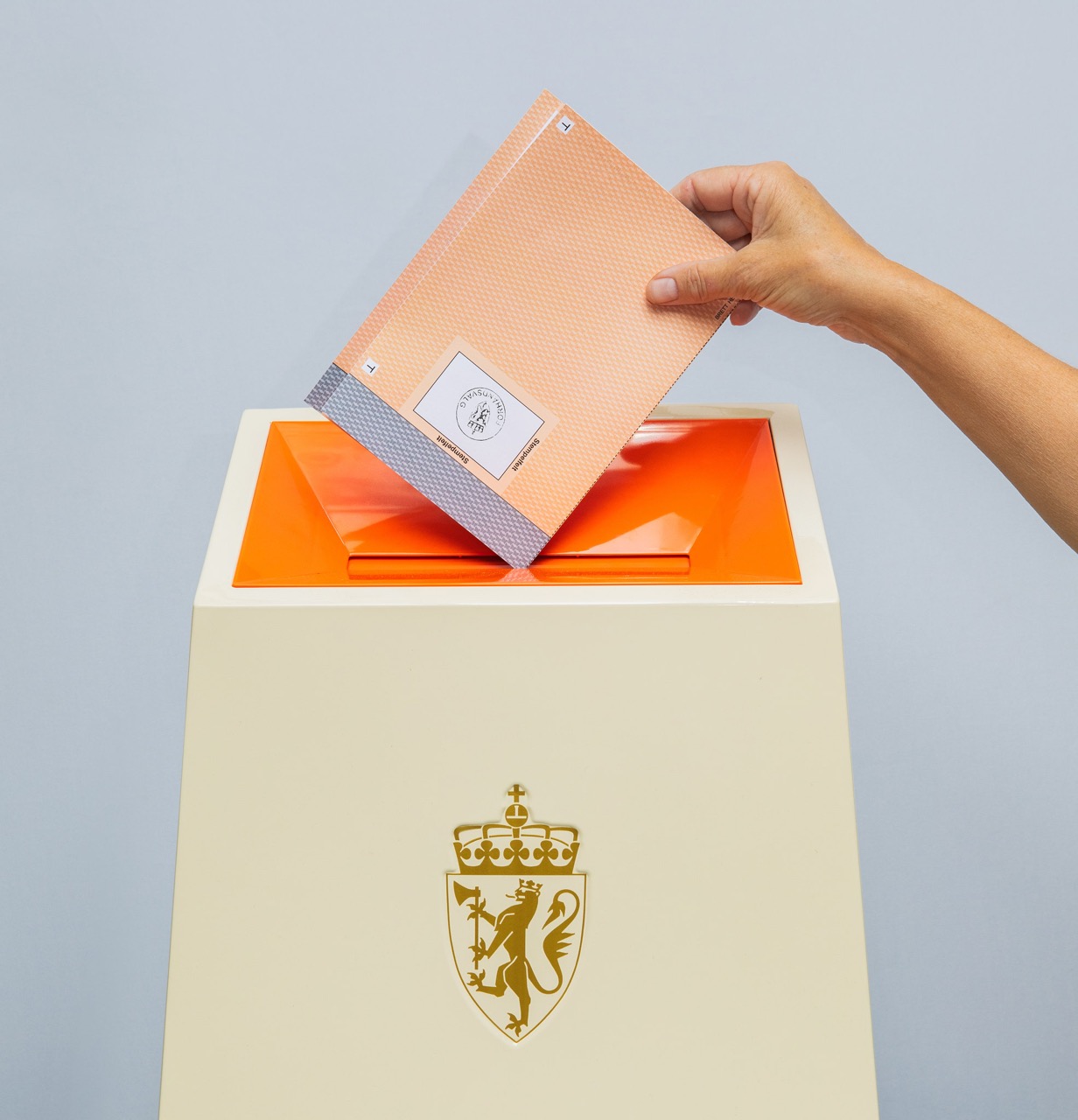 Illustrasjonsbilde av en hånd som legger en stemmeseddel i en valgurne merket med riksvåpenet. Bildet symboliserer stemmegivning ved stortingsvalget 2025